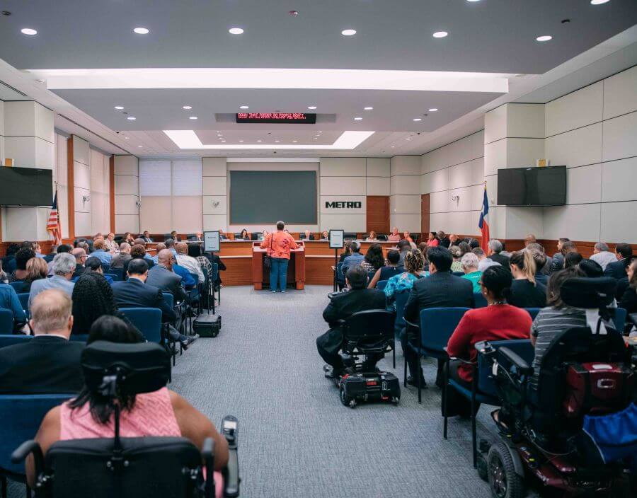 View from the back of the METRO boardroom during a meeting of the Board of Directors with many people in attendance including some in motorized wheelchairs.